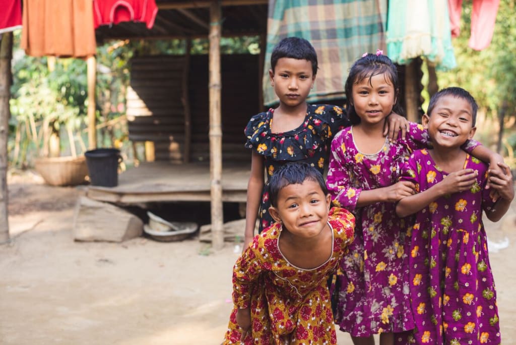 Four little girls wearing colourful dresses laugh and pose together in front of their home.